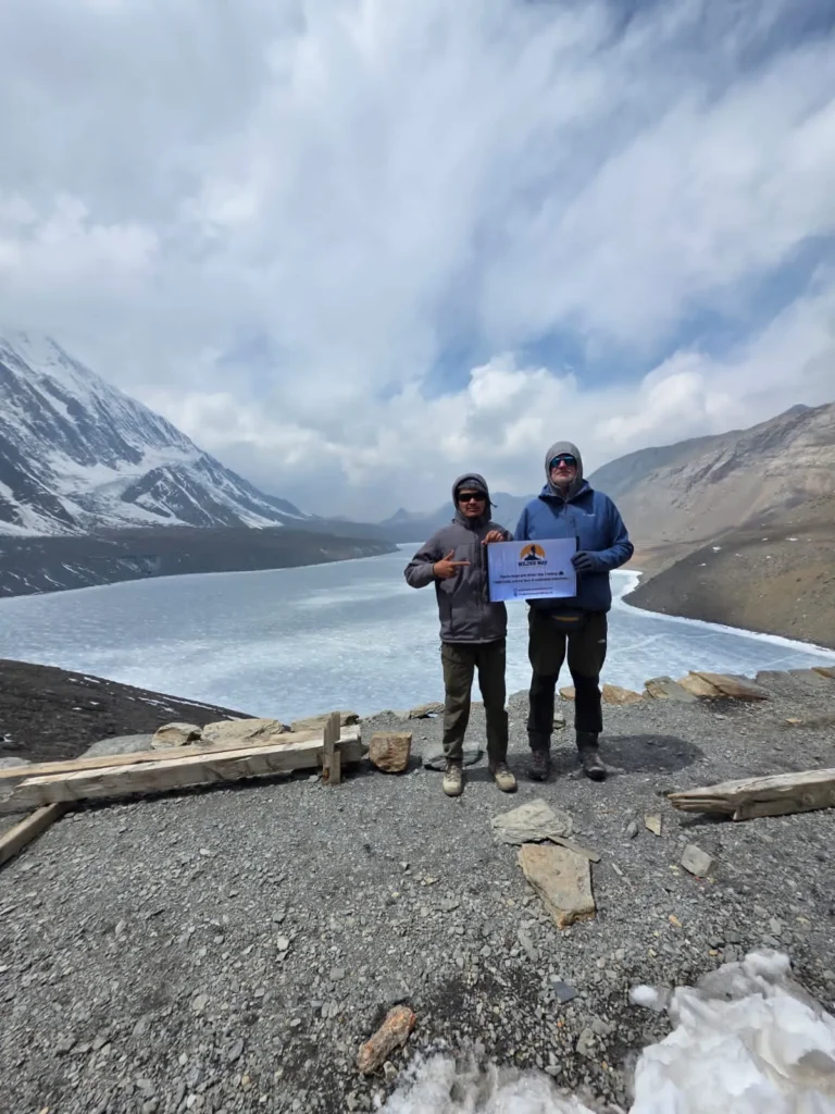 Two trekkers overlooking the vast frozen expanse of Tilicho Lake under a cloudy sky on the Annapurna Circuit Trek with Tilicho Lake.