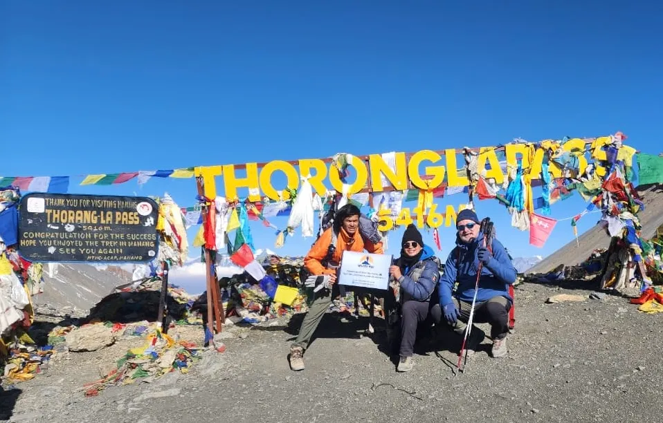 Trekkers celebrating at the yellow Thorong La Pass sign (5416m), the highest point of the Annapurna Circuit Trek with Tilicho Lake.